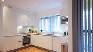 Une cuisine moderne avec des meubles blancs, un four encastré, un four à micro-ondes et une fenêtre équipée de stores.