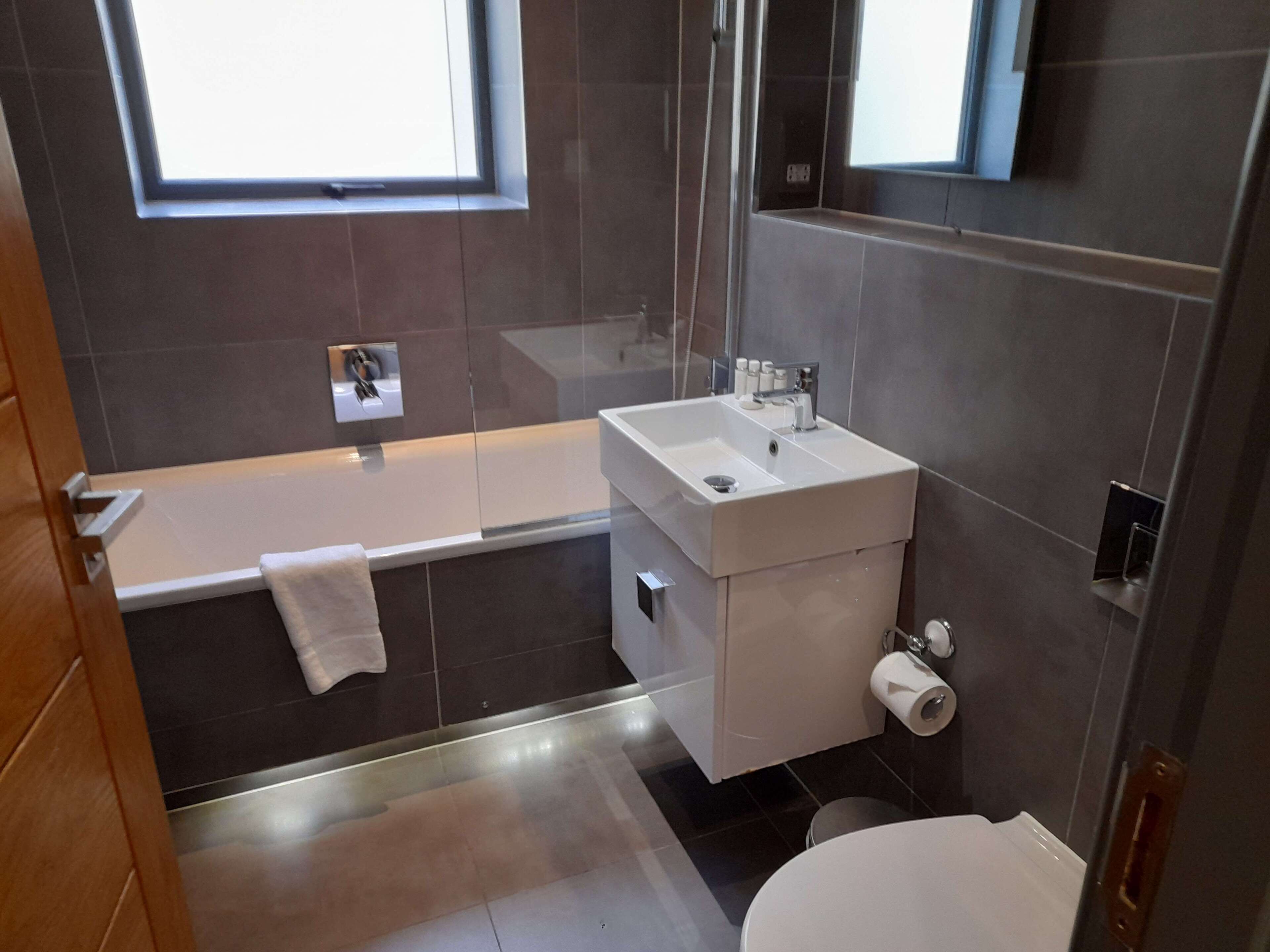 A modern bathroom with a bathtub, sink, and toilet. There are two windows providing natural light.