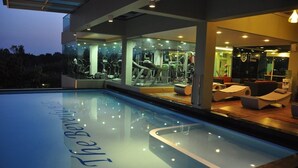 Un espace piscine intérieur moderne avec un mur en verre, des équipements de salle de sport et des fauteuils confortables.