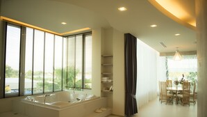 Une salle de bain moderne avec une grande baignoire, un lavabo et une douche. On y trouve des fenêtres équipées de stores, un coin repas avec une table et des chaises, ainsi qu’un lustre.