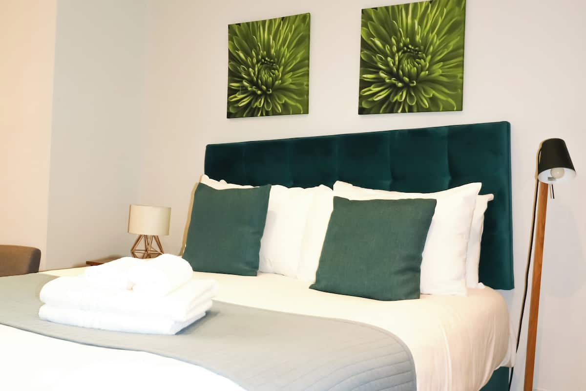 Un lit bien fait, avec une tête de lit de couleur turquoise, une literie blanche et des coussins verts. Deux tableaux représentant des motifs floraux verts sont accrochés au mur au-dessus du lit.