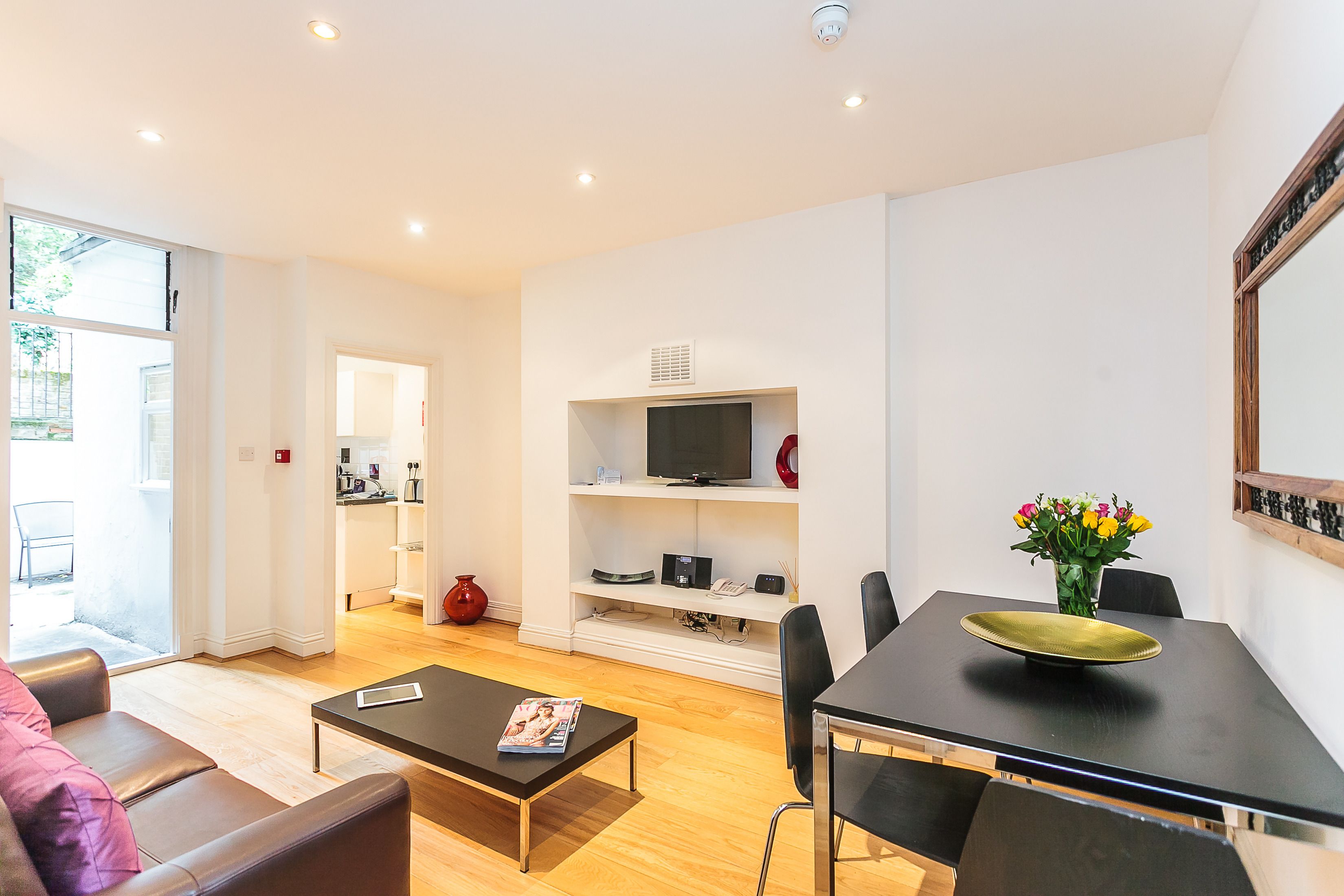 Una sala de estar moderna con un sofá, una mesa de centro y una mesa de comedor con sillas. Hay un televisor en un estante, un florero con flores y un florero rojo en el suelo.