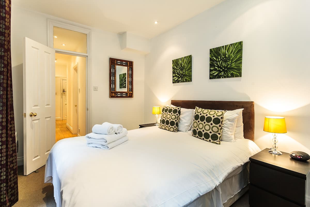 Un lit bien fait, avec du linge de lit blanc et des coussins décoratifs, une lampe de chevet et un miroir au mur.
