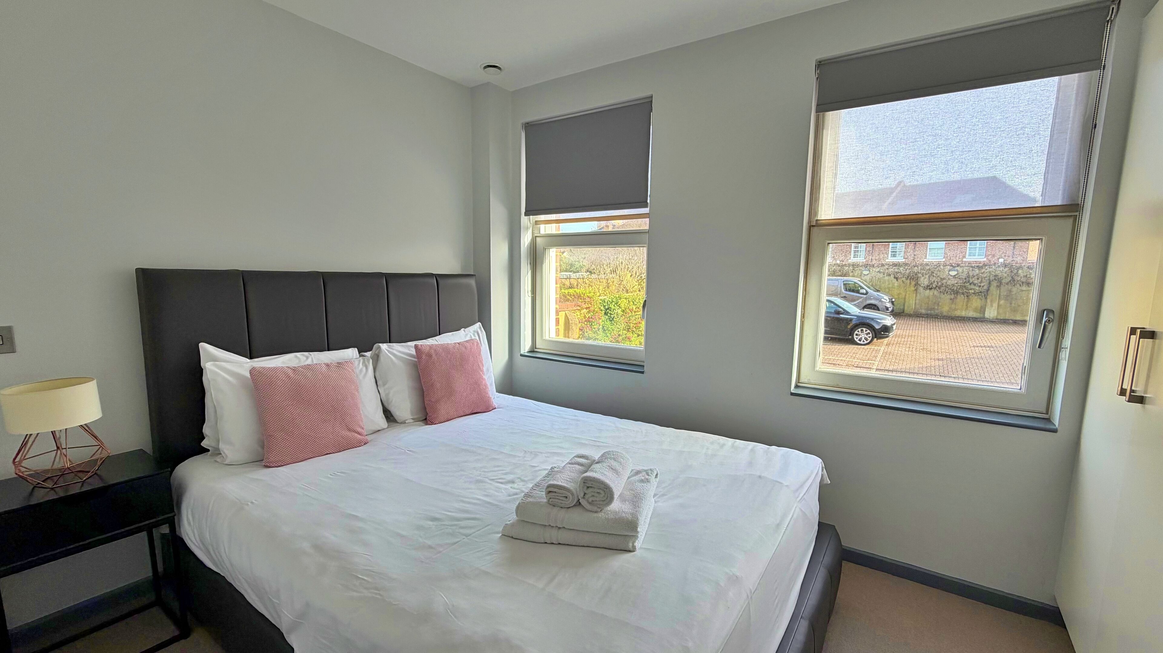 Une chambre d’hôtel avec un grand lit, deux fenêtres équipées de stores, une table de chevet avec une lampe, et une vue sur une voiture garée à l’extérieur.