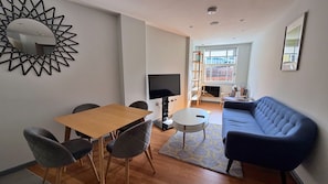 Uma sala de estar moderna com uma mesa de jantar de madeira, cadeiras estofadas cinzentas, um sofá azul, uma TV de tela plana e um espelho decorativo.