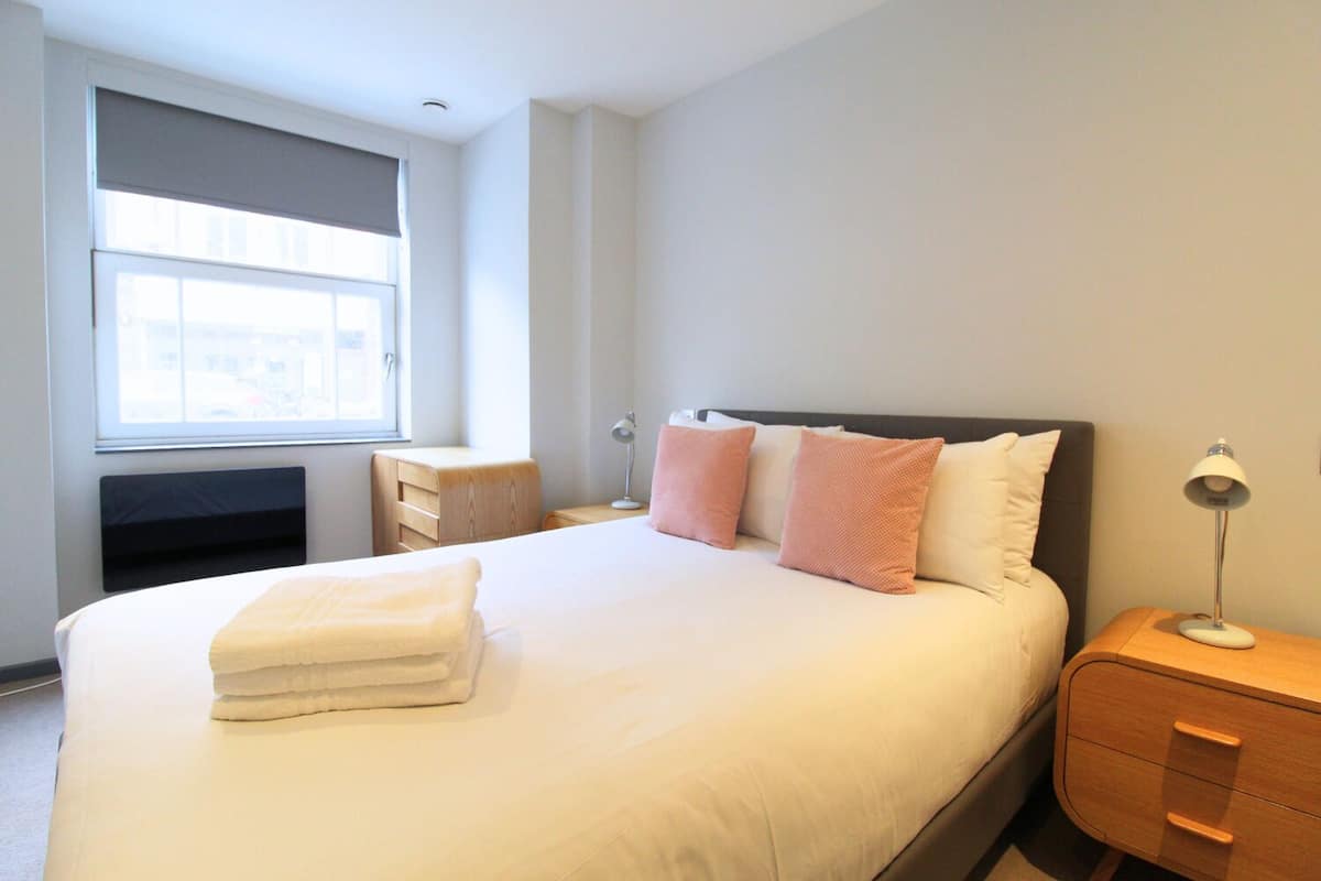 A neatly made bed with a folded towel, a bedside table with a lamp, and a window with blinds.