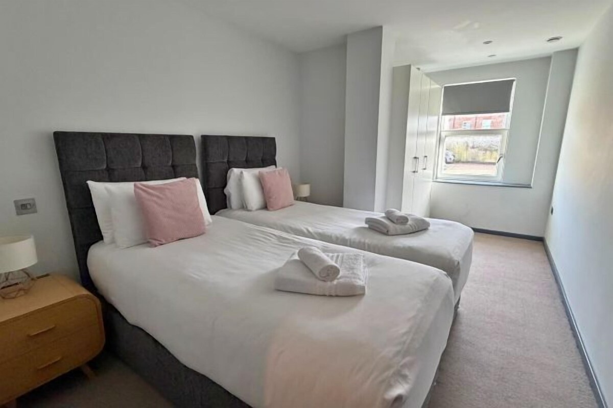 Quarto de hotel com duas camas, uma mesinha de cabeceira e uma janela.