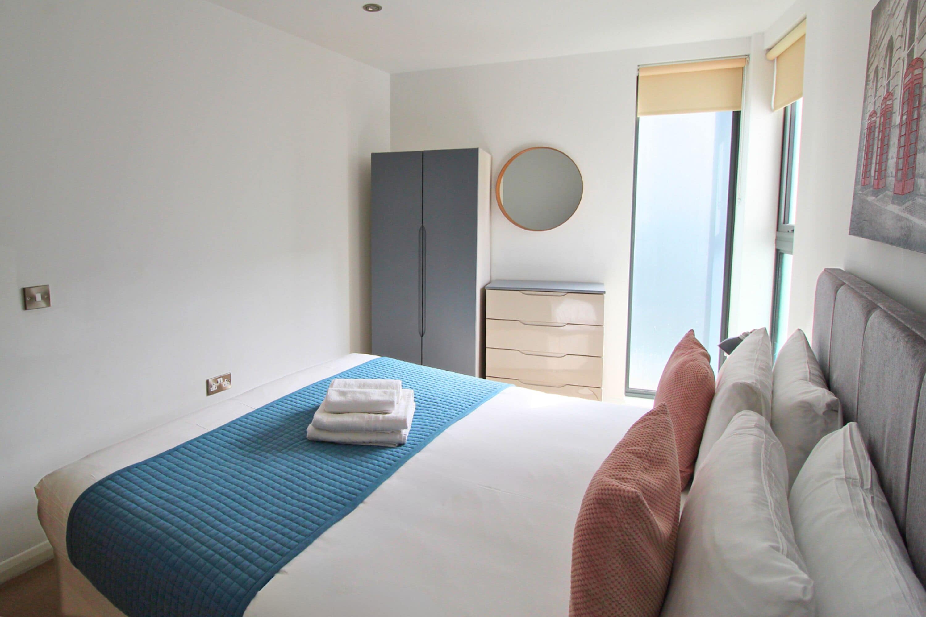 A bedroom with a bed, a wardrobe, a dresser, a round mirror, and a window.