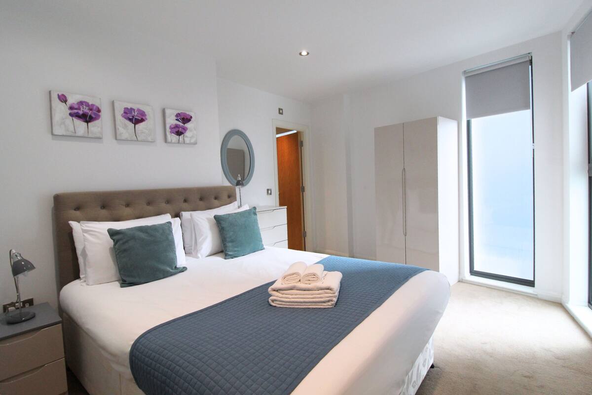 Une chambre à coucher moderne avec un grand lit, une table de chevet, un miroir et des œuvres d’art accrochées au mur.
