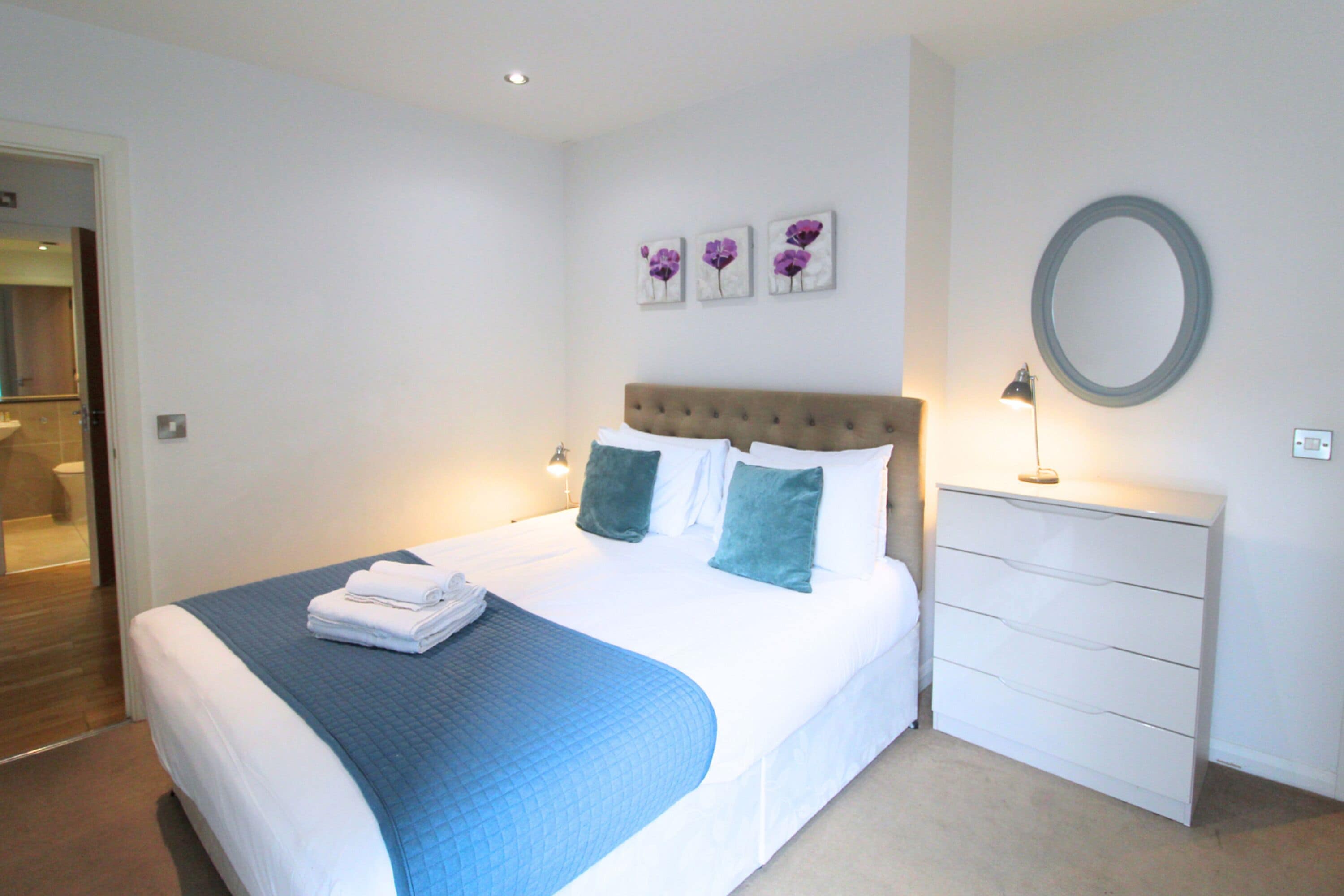 Een net opgemaakt bed met een blauwe deken, een nachtkastje met een lamp, en een spiegel aan de muur.