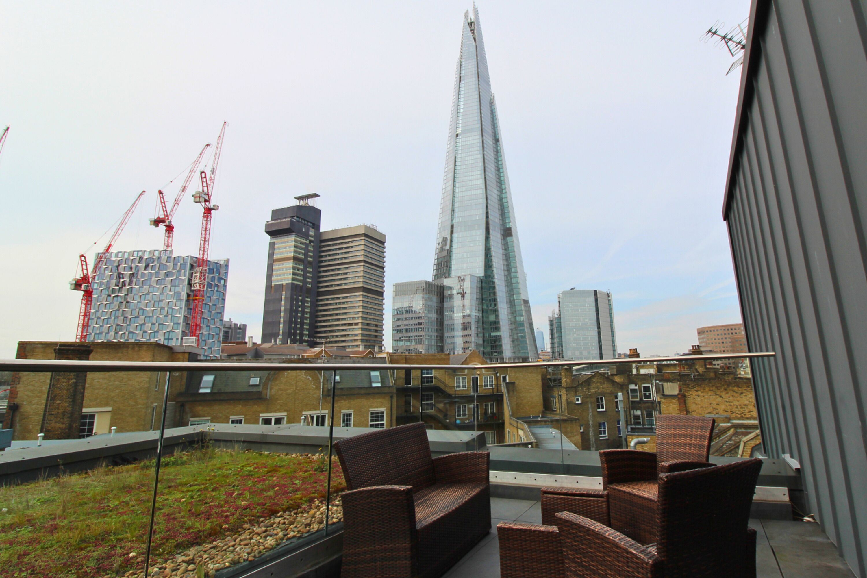A rooftop terrace with wicker furniture, a view of the city skyline including the Shard, and construction cranes in the background.