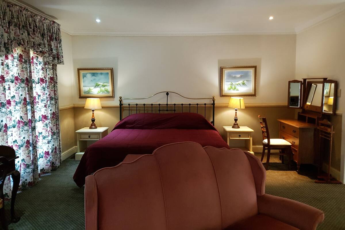 Habitación de hotel con una cama, dos mesitas de noche con lámparas, una silla, un tocador con espejo y cortinas con estampado floral.