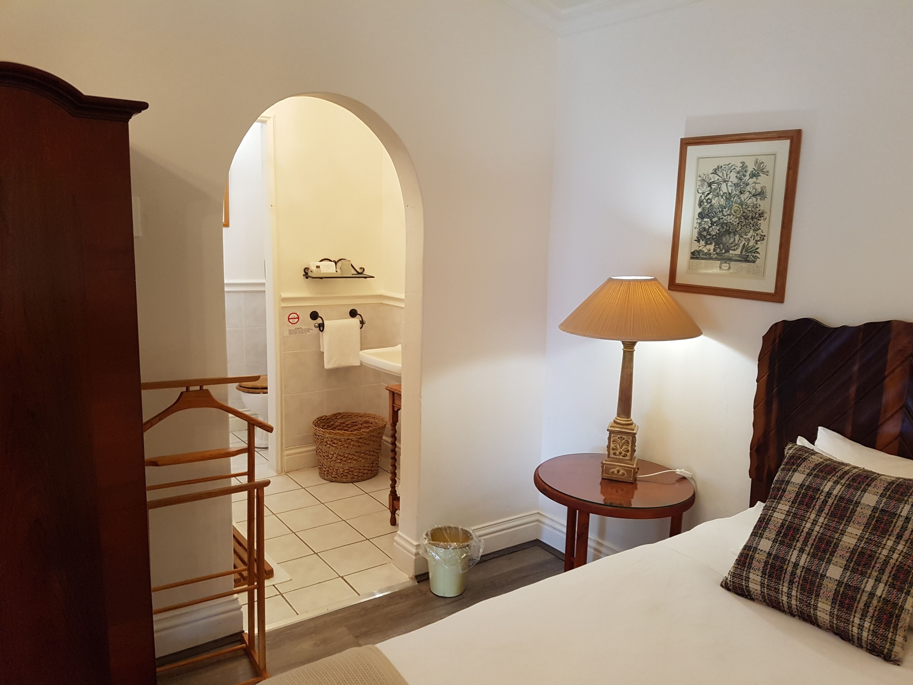 A hotel room with a bed, a nightstand, a lamp, a framed picture, a wooden headboard, a small table, a basket, and a towel rack.