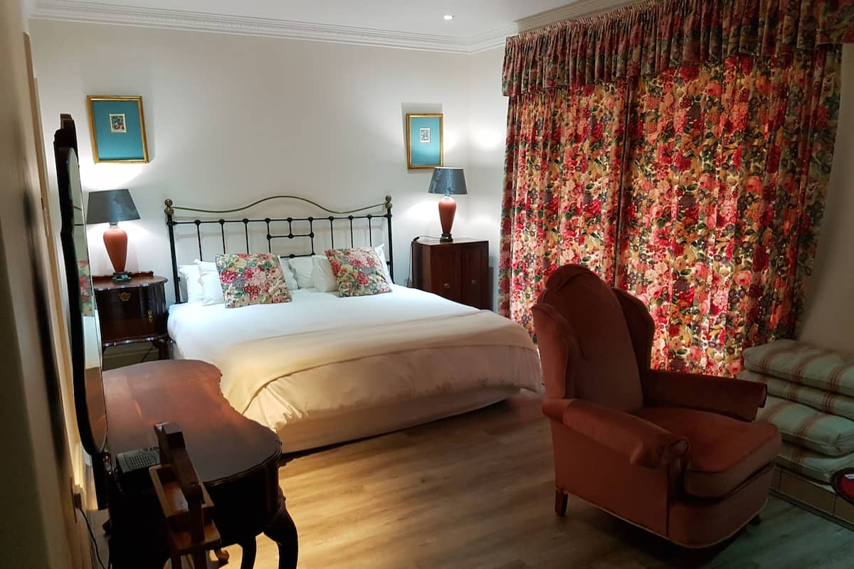 Un dormitorio con cama, dos mesitas de noche, una silla, un escritorio y un ventanal con cortinas florales.