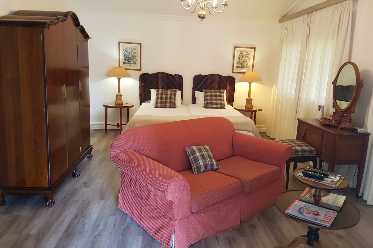 Una habitación de hotel con una cama grande, dos mesitas de noche con lámparas, un armario de madera, un sofá rojo, una mesa de centro redonda con revistas y una araña.