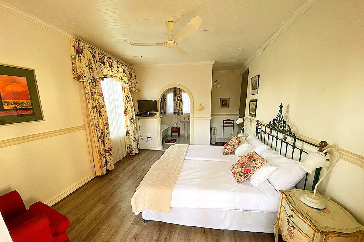 Una habitación de hotel con una cama, una silla roja, una mesita de noche, un ventilador de techo y un cuadro en la pared.