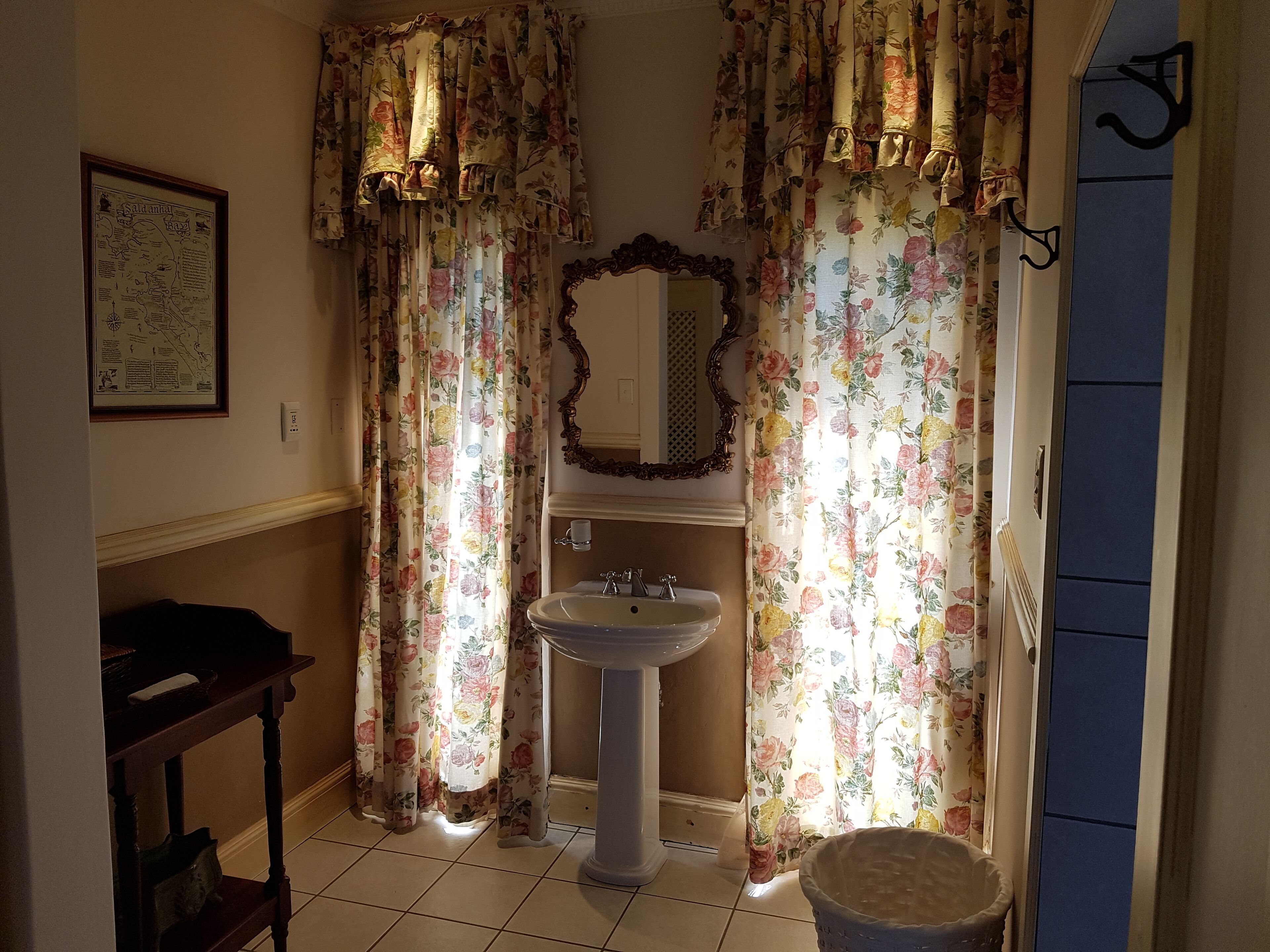 Une salle de bain avec des rideaux à motifs floraux, un lavabo sur pied, un miroir et un tableau encadré accroché au mur.