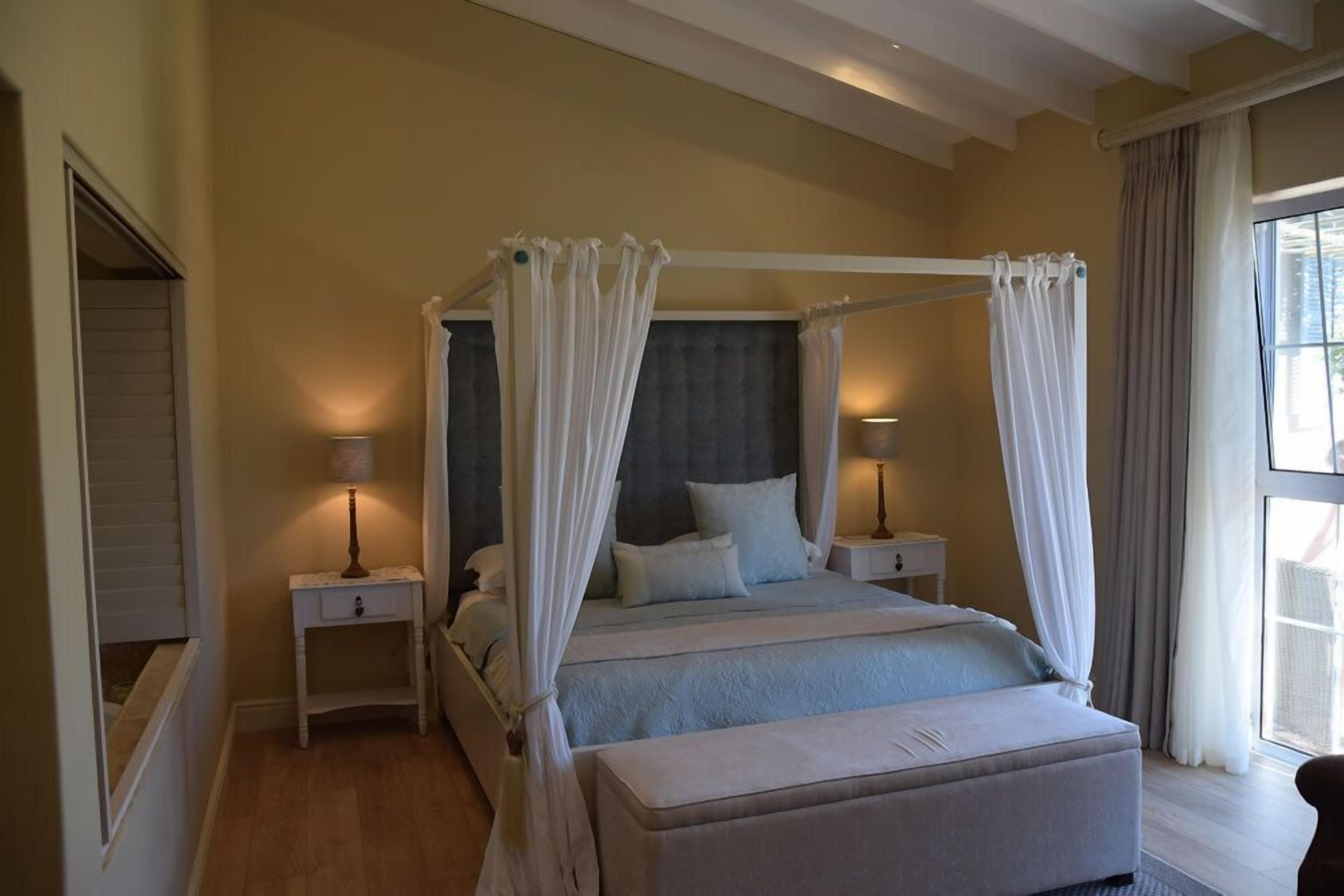 Una cama con dosel y cortinas blancas, una mesita de noche con lámpara y un ventanal con cortinas.