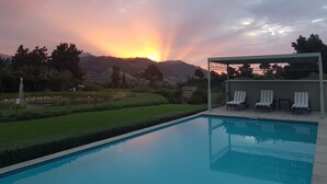 A swimming pool with a pergola and lounge chairs, surrounded by a landscaped garden and a view of a sunset over hills.