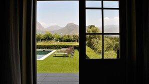 A view from a room with a large window overlooking a well-maintained garden with a swimming pool, a wooden deck, and distant mountains.