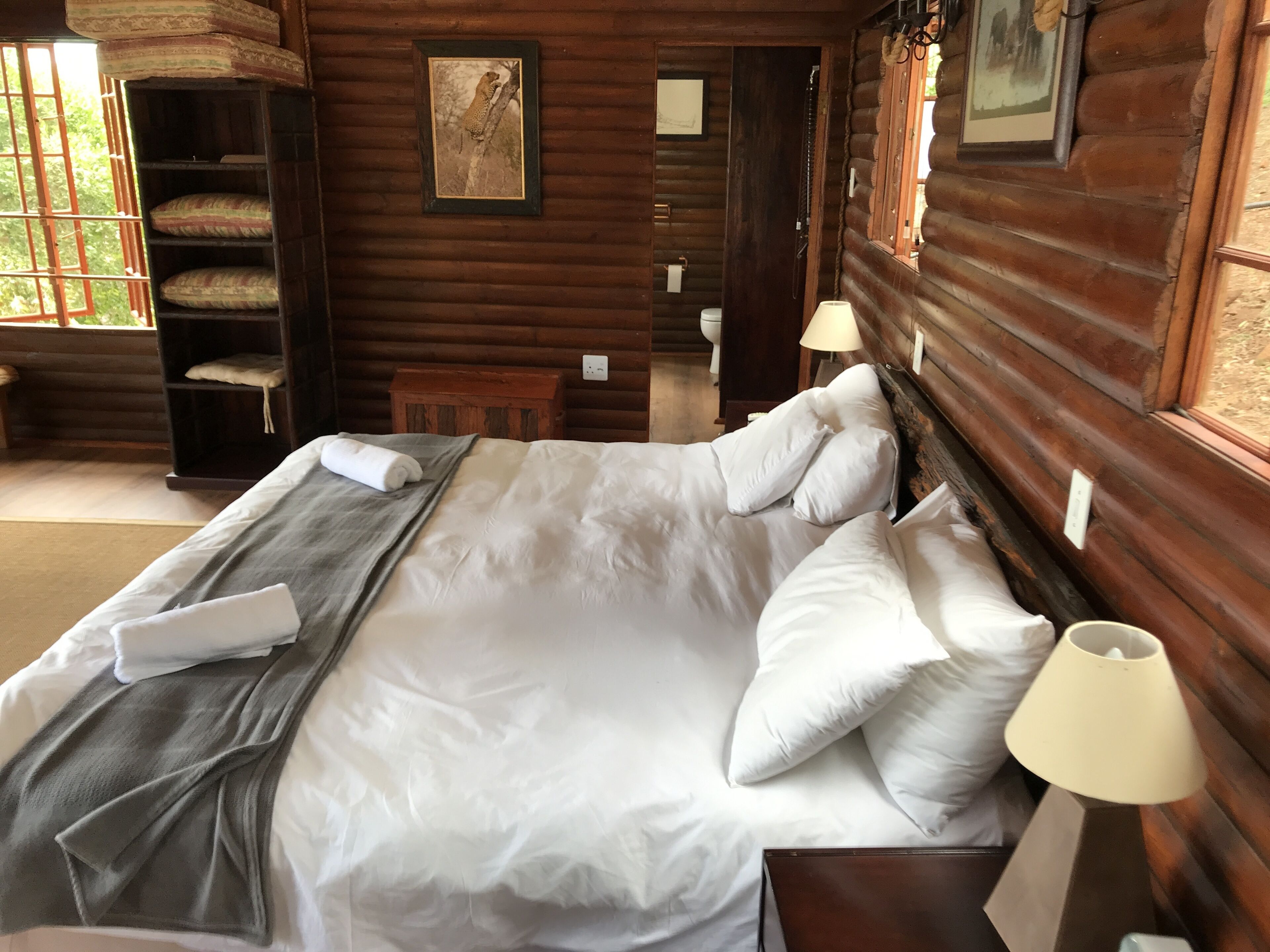 A wooden cabin bedroom with a large bed, pillows, a bedside lamp, a framed picture on the wall, and a window with a view of trees.