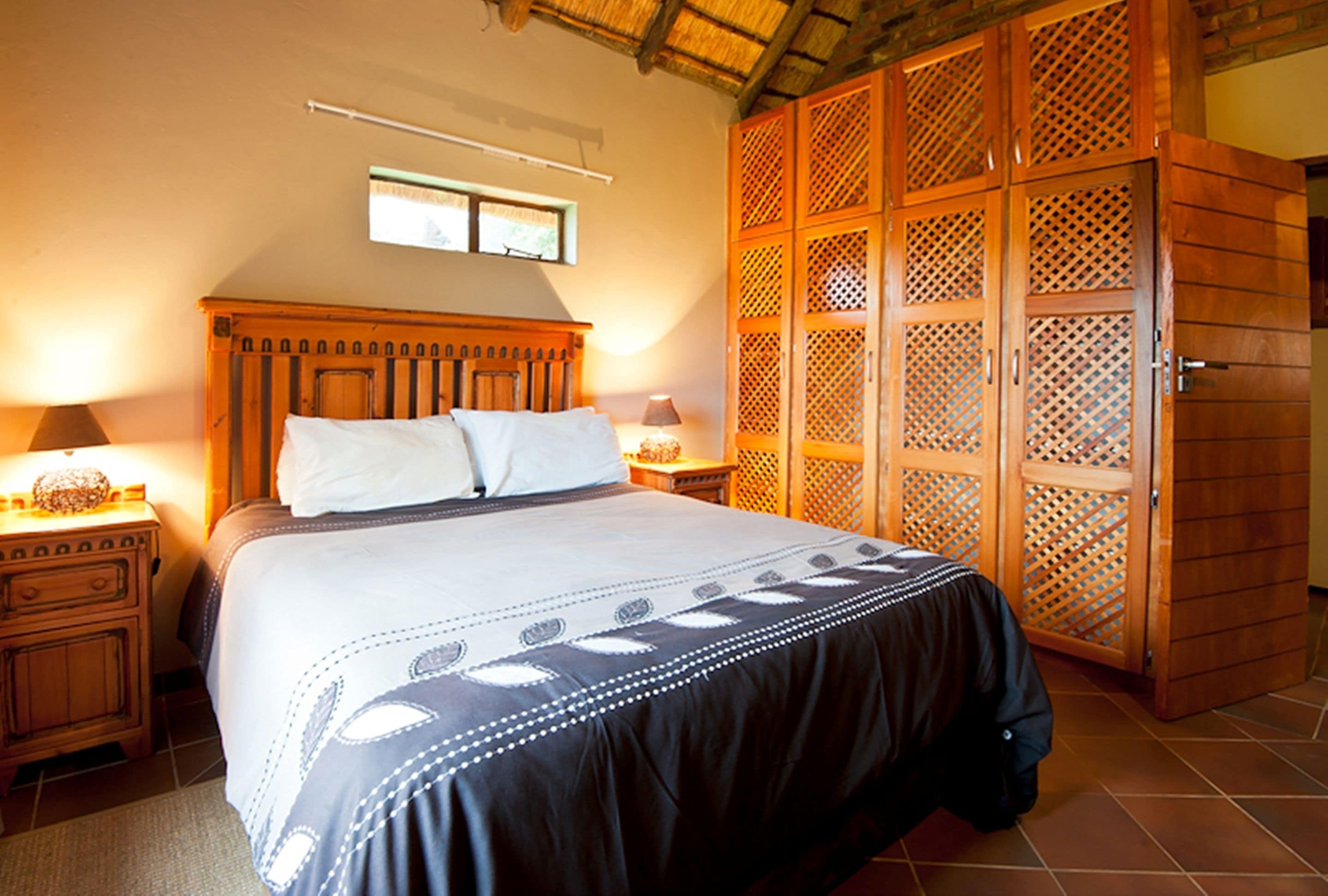Un dormitorio con un marco de cama de madera, dos mesitas de noche con lámparas, un ventanal grande y un armario de madera con diseño de celosía.