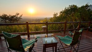 Una terraza de madera con dos sillas plegables verdes y una mesita, con vistas a un paisaje pintoresco con árboles y un atardecer.
