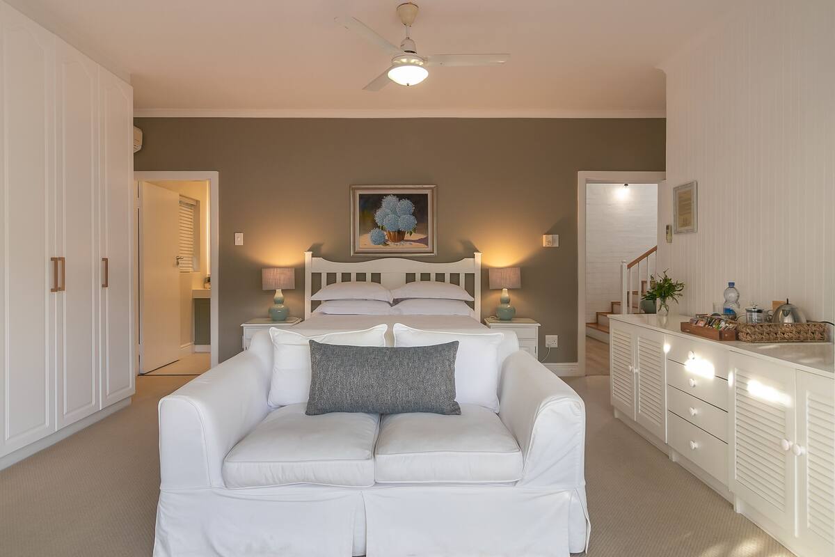 A bedroom with a bed, bedside tables, a white wardrobe, a white sofa with a pillow, and a ceiling fan.