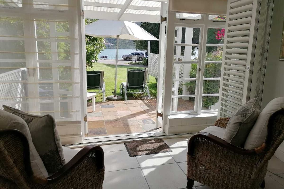 A room with a view of a patio and a body of water. There are two wicker chairs and a small table.