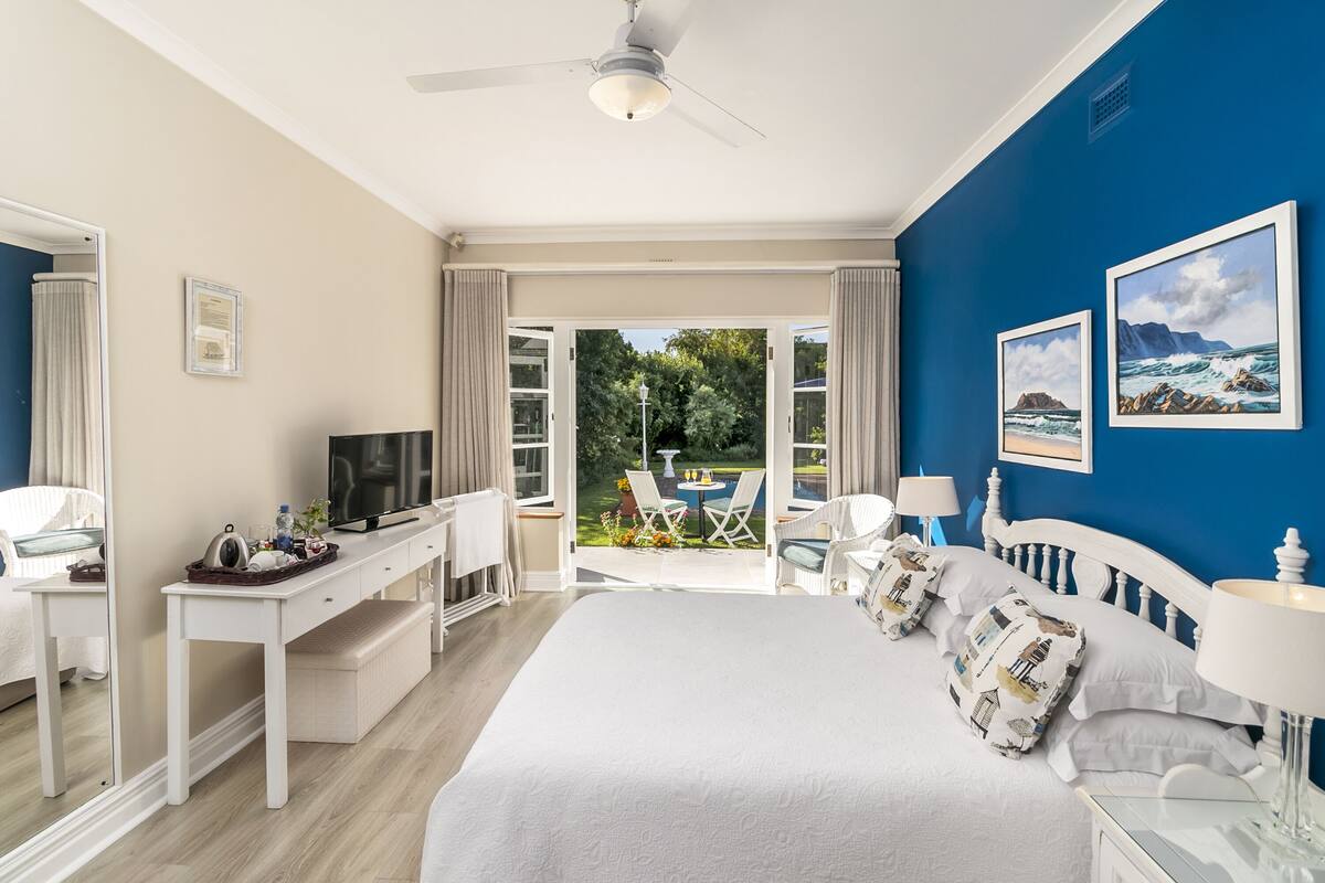 A bedroom with a large bed, a desk, a TV, and a view of the garden.