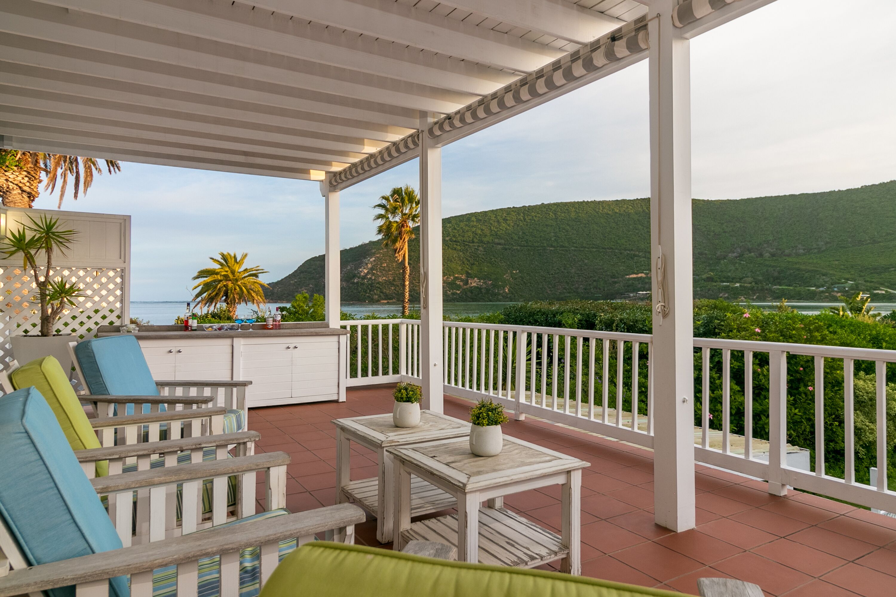 A covered outdoor area with white furniture, a small table with potted plants, and a view of a green landscape and hills.