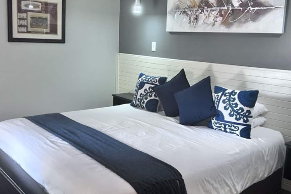 A neatly made bed with a white and blue bedspread and matching pillows, a framed picture on the wall, and a modern pendant light.