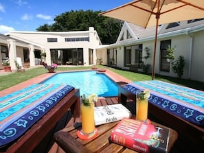 Ein Poolbereich mit Holzterrasse, überdachter Sitzgelegenheit und einem Tisch, auf dem das Buch 'Monsoon' liegt.