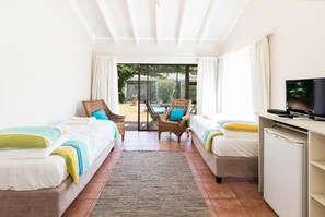 A hotel room with two single beds, a TV, a desk, and a view of a pool and garden.