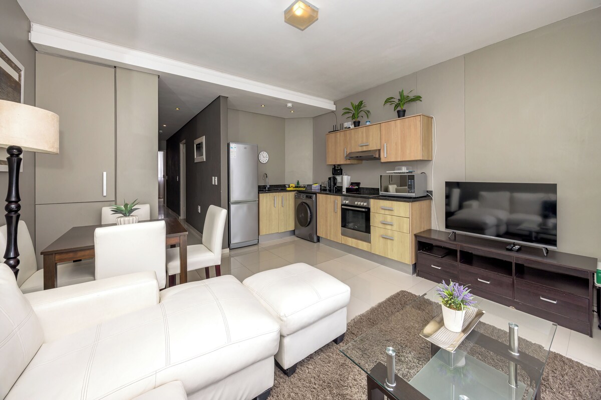A modern living room with a sofa, a glass coffee table, a TV, and a kitchen area with cabinets and appliances.