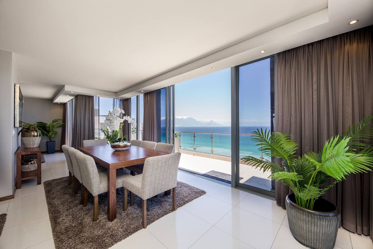 A modern dining area with a wooden table and chairs, a large potted plant, and a view of the ocean through floor-to-ceiling windows.