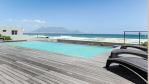 A wooden deck with a pool, beach view, and a mountain in the background. - Infinity Self-Catering Apartments (Cape Town)