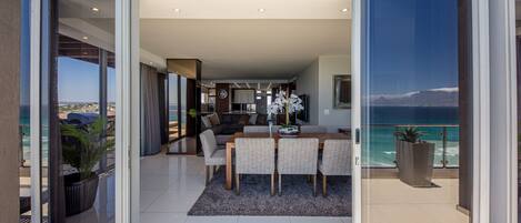 A modern living room with a dining area, large sliding glass doors leading to a balcony with a view of the ocean.