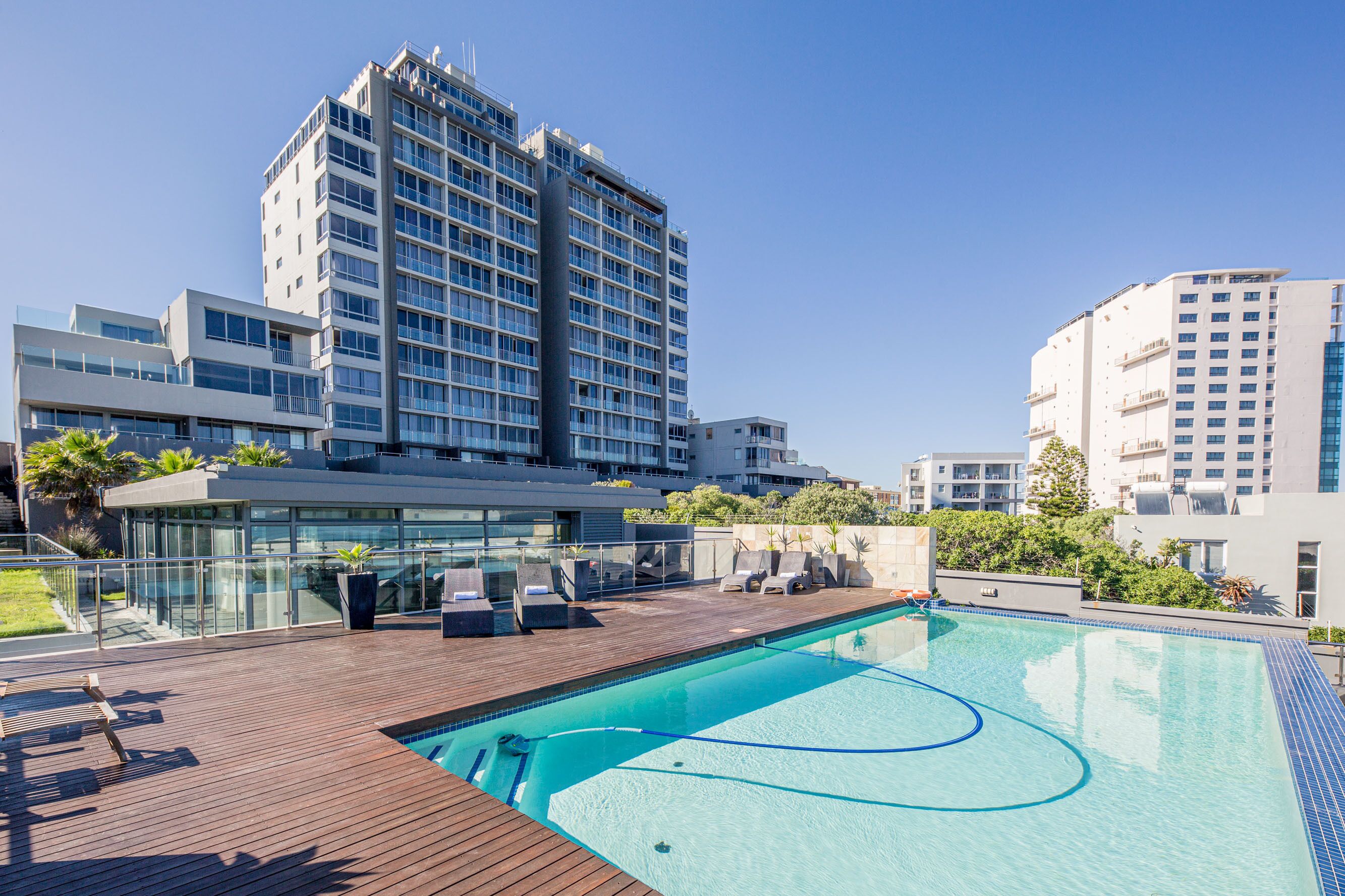 Una piscina en la azotea con sillones de descanso, barandilla de cristal y edificios modernos al fondo.