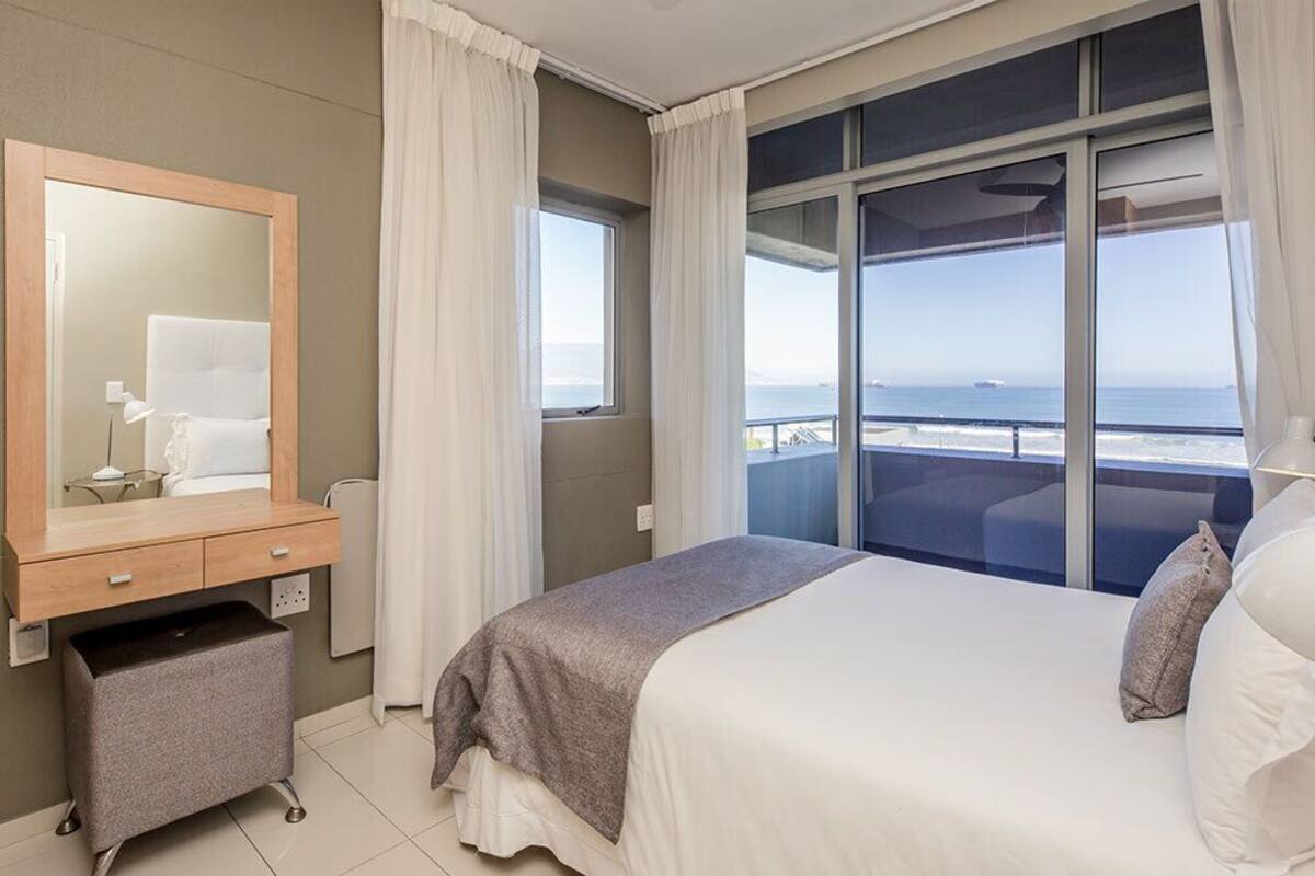 A hotel room with a bed, a dresser, a mirror, a window with a view of the sea, and a small desk with a lamp.