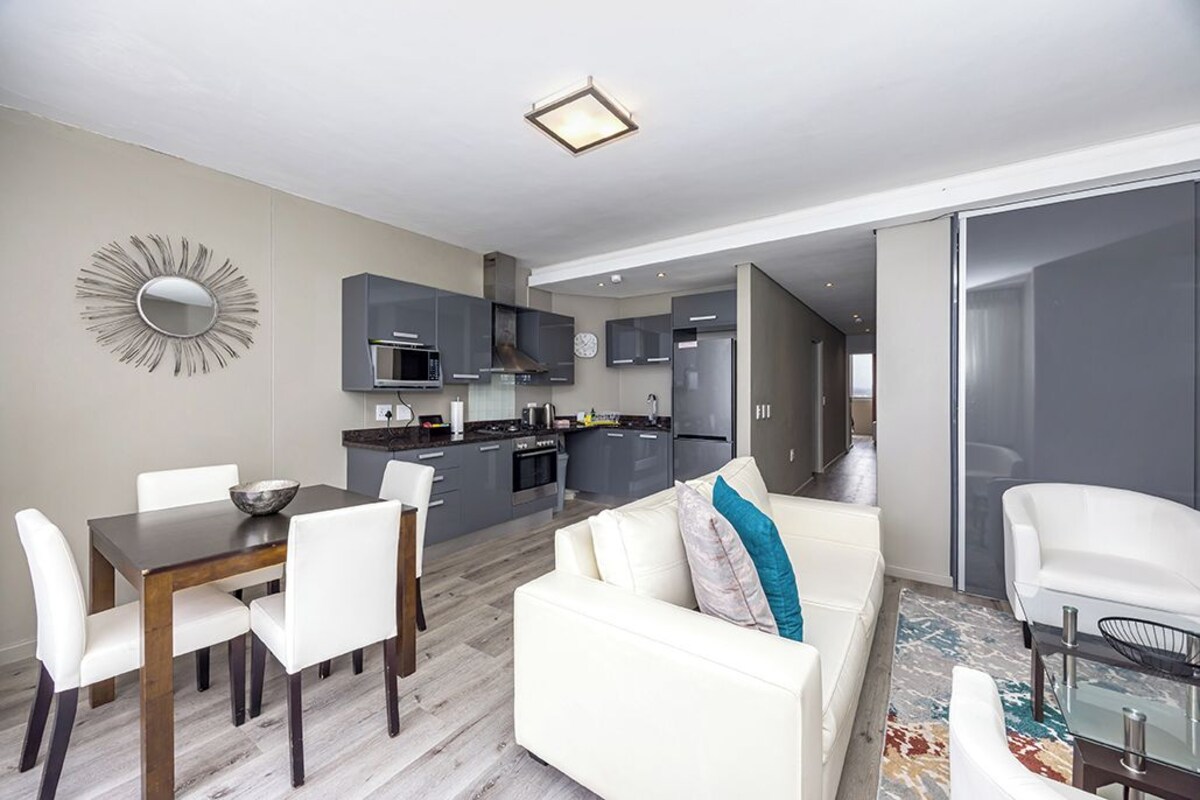 A modern kitchen with grey cabinets, a dining table with white chairs, and a white sofa with a blue pillow.