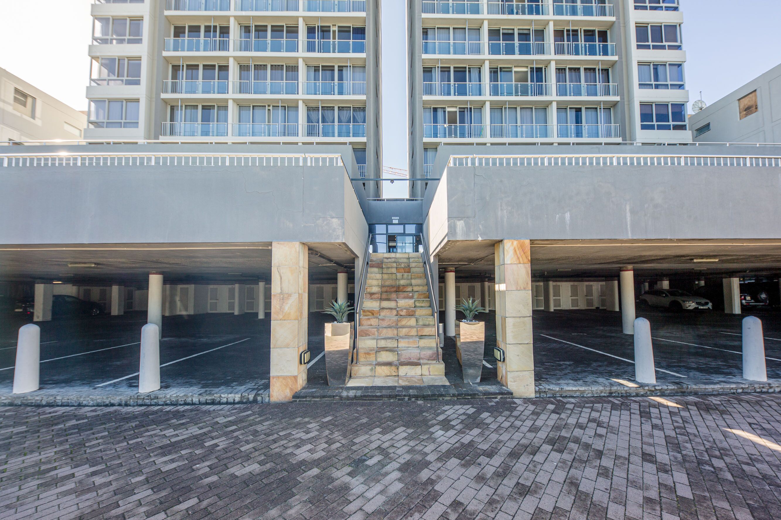 A parking garage entrance with a central staircase leading to upper levels, surrounded by parking spaces and bollards.