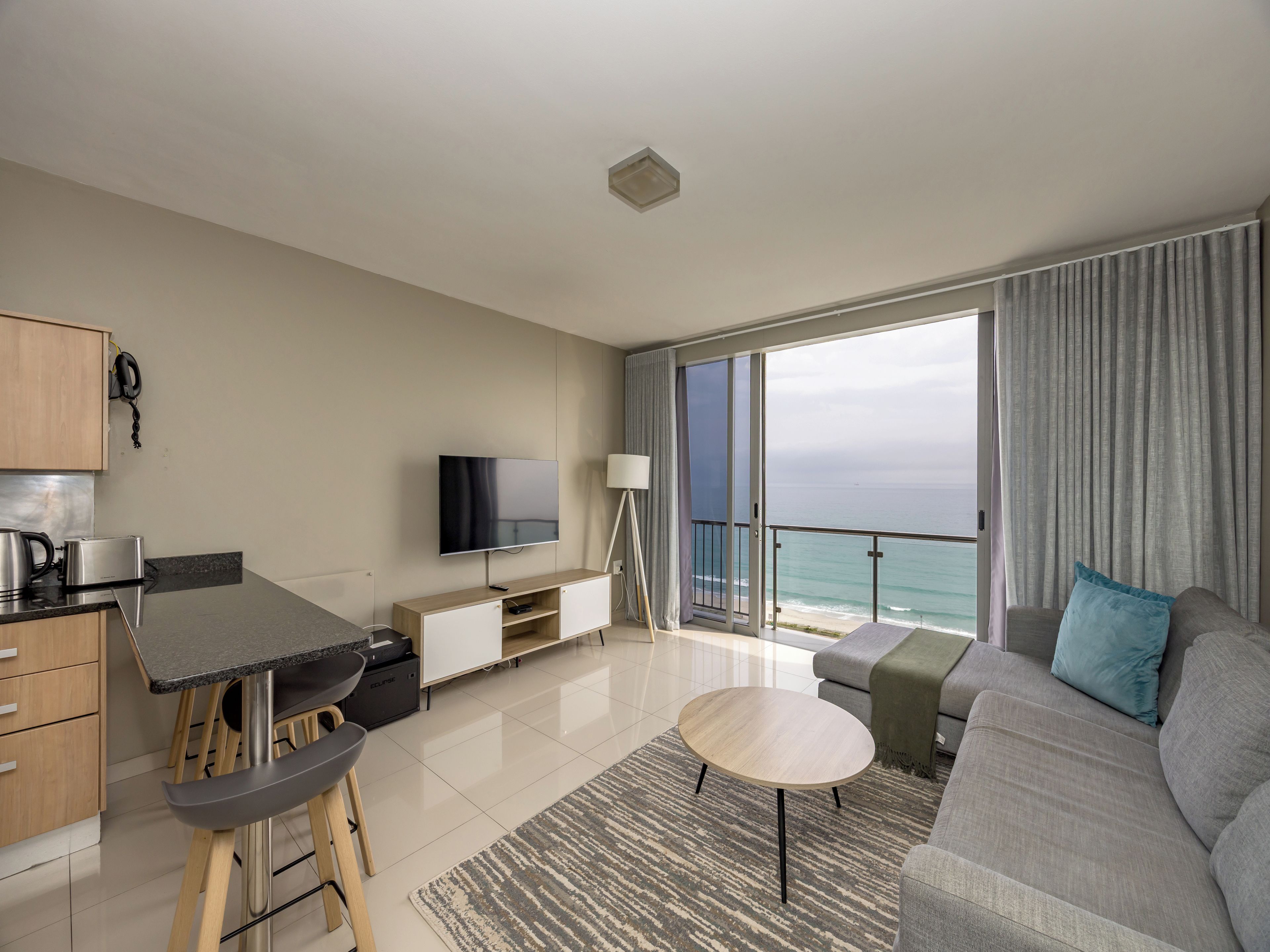 A modern living room with a flat-screen TV, a dining area with a bar stool, a sofa with a throw pillow, and a view of the ocean through large windows.