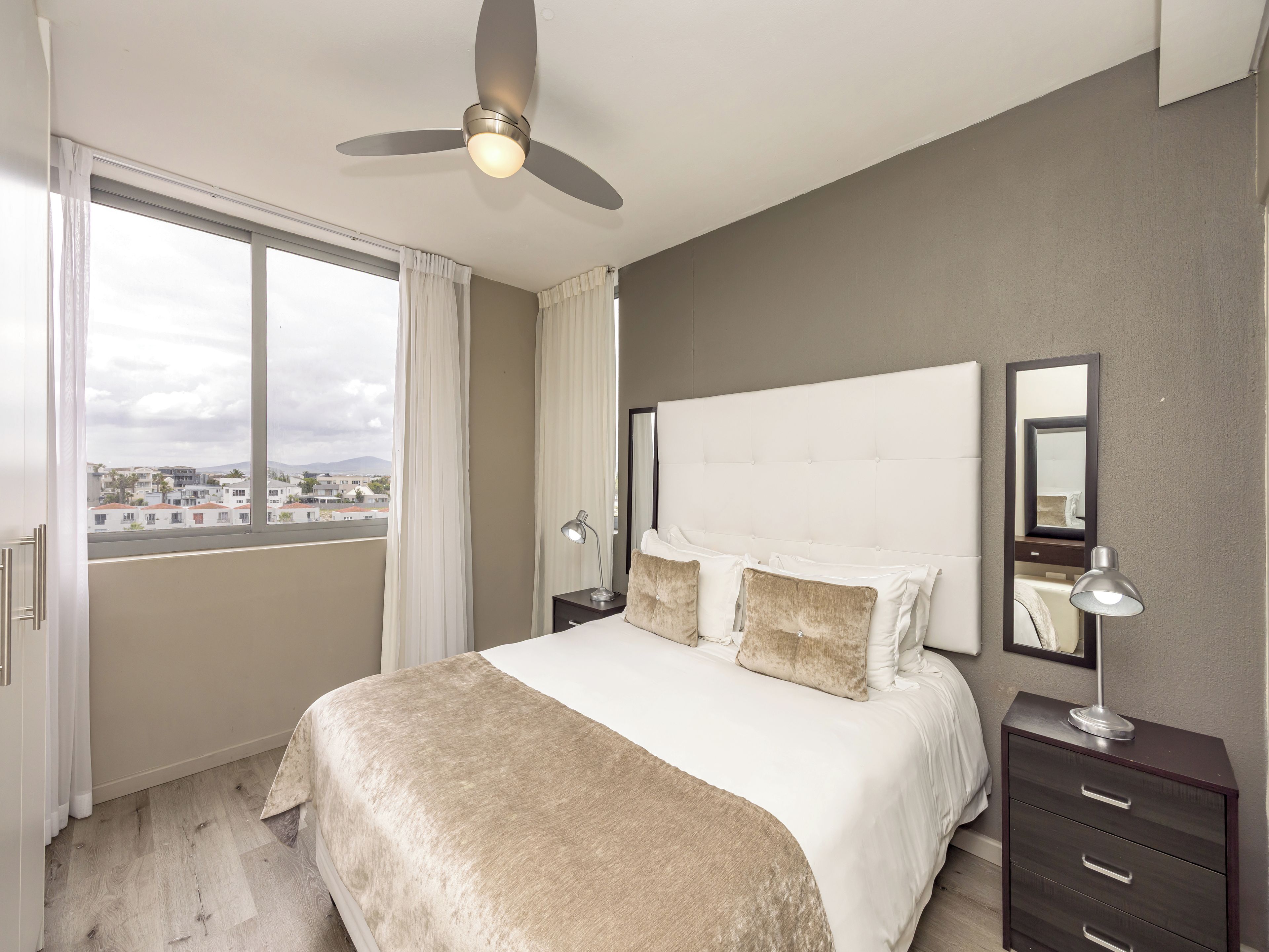 A hotel room with a large bed, a ceiling fan, a nightstand with a lamp, a dresser, and a view of the city through the window.