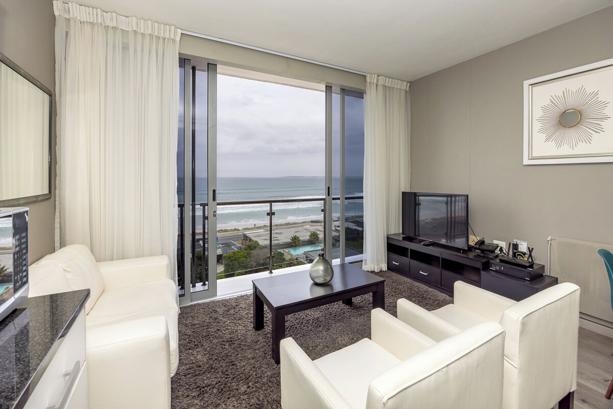 A modern living room with a large window, a flat-screen TV, and a view of the ocean.