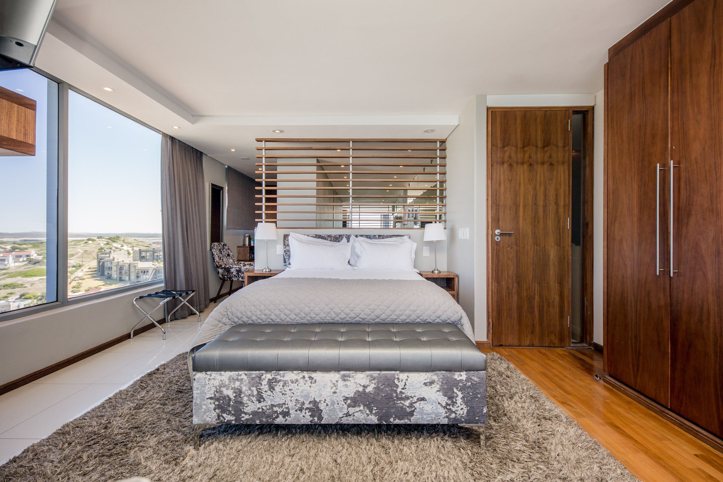 A modern bedroom with a large bed, a view of the city, and a wardrobe.