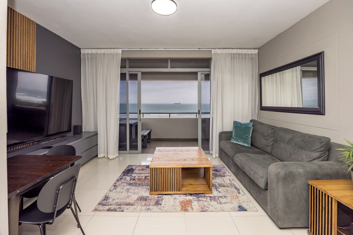 A modern living room with a flat-screen TV, a grey sofa, a wooden coffee table, and a view of the ocean through large windows.