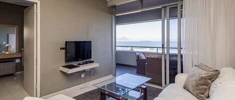 A modern living room with a glass coffee table, white sofa, and a flat-screen TV on a shelf. A balcony with a view of the sea and a mountain is visible through the glass doors.