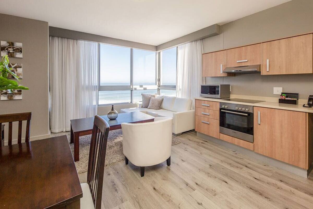 A modern kitchen with wooden cabinets, a microwave, and an oven. There is a dining area with a table and chairs, and a sofa. The room has large windows with a view of the ocean.