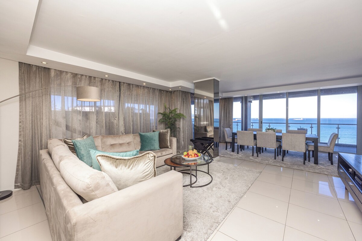 A modern living room with a sofa, coffee table, and a dining area with chairs and a view of the sea.